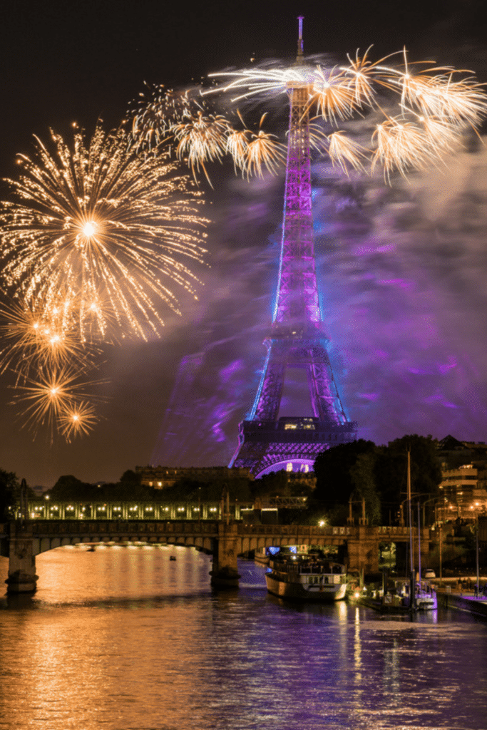 The Eiffel Tower glowing with purple and blue lights, surrounded by golden fireworks, with the Seine River and a nearby bridge illuminated at night.