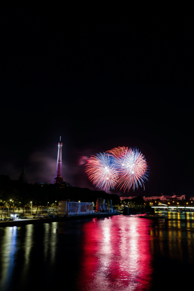 The Eiffel Tower lit up in red and blue lights, with large red and blue fireworks bursting in the sky, reflecting on the Seine River at night.