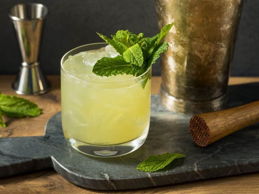 A refreshing green Chartreuse cocktail garnished with a sprig of fresh mint, accompanied by bar tools on a slate surface.