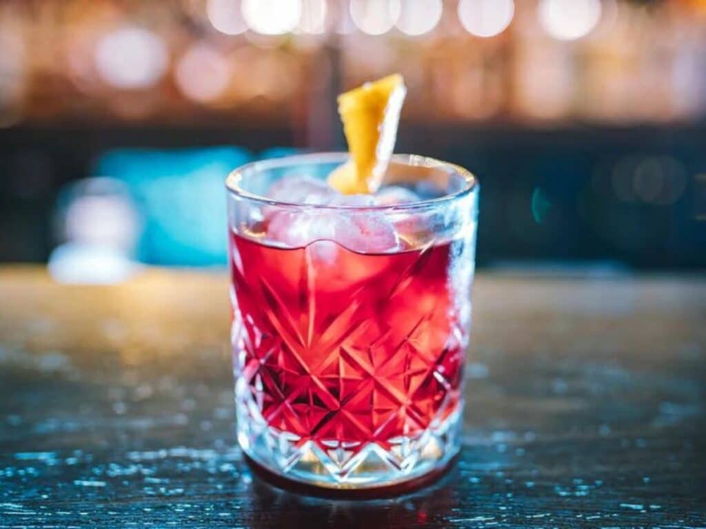 A vibrant red Dubonnet cocktail with ice cubes, garnished with a twist of lemon peel, in a patterned glass against a blurred bar background.