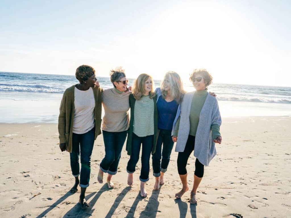 A group of women walking arm in arm on a sunny beach, enjoying each other's company.