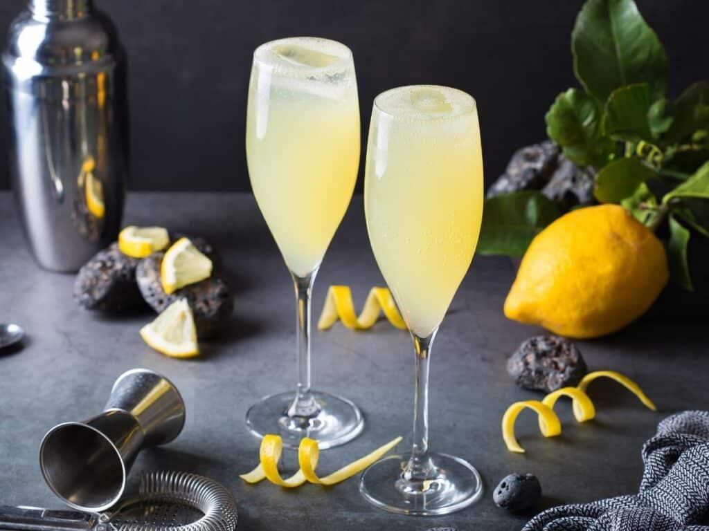 Two elegant French 75 cocktails in champagne flutes, garnished with lemon twists, accompanied by cocktail-making tools and fresh lemons on a dark surface.