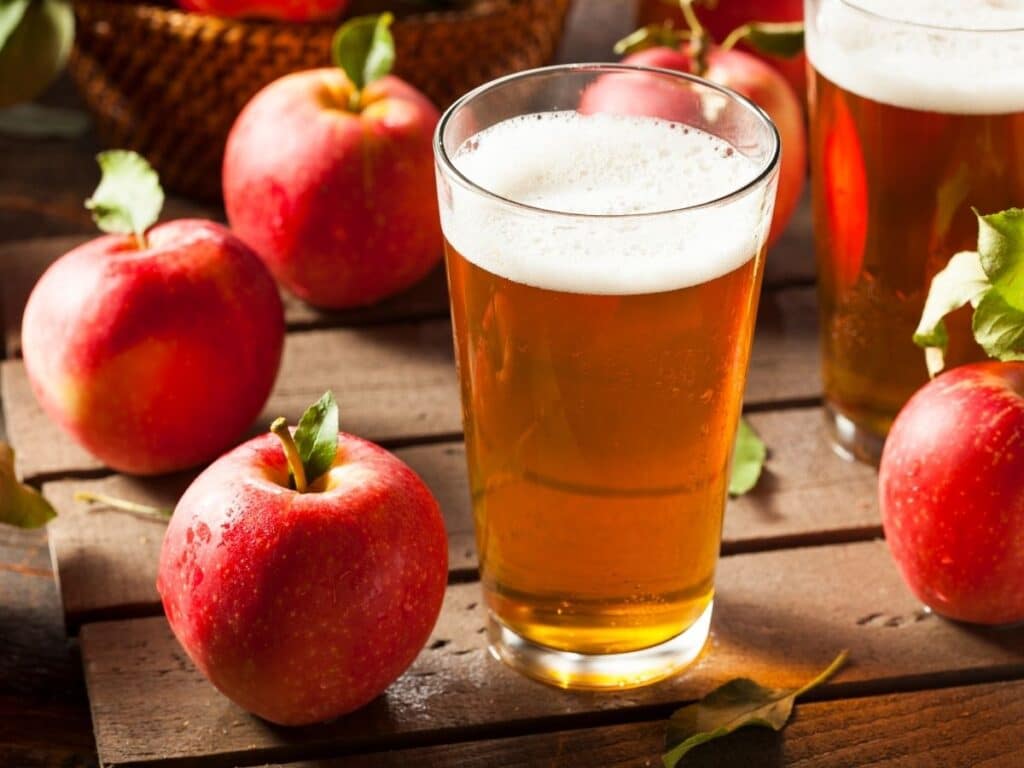 A glass of French apple cider with a foamy head, surrounded by fresh red apples, set on a rustic wooden surface.