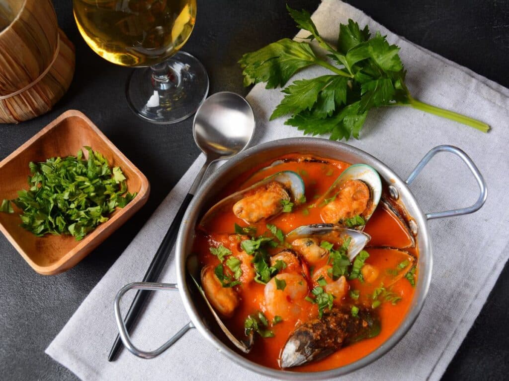 A bowl of bouillabaisse, a traditional French seafood stew, garnished with fresh herbs, with a glass of white wine on the side.