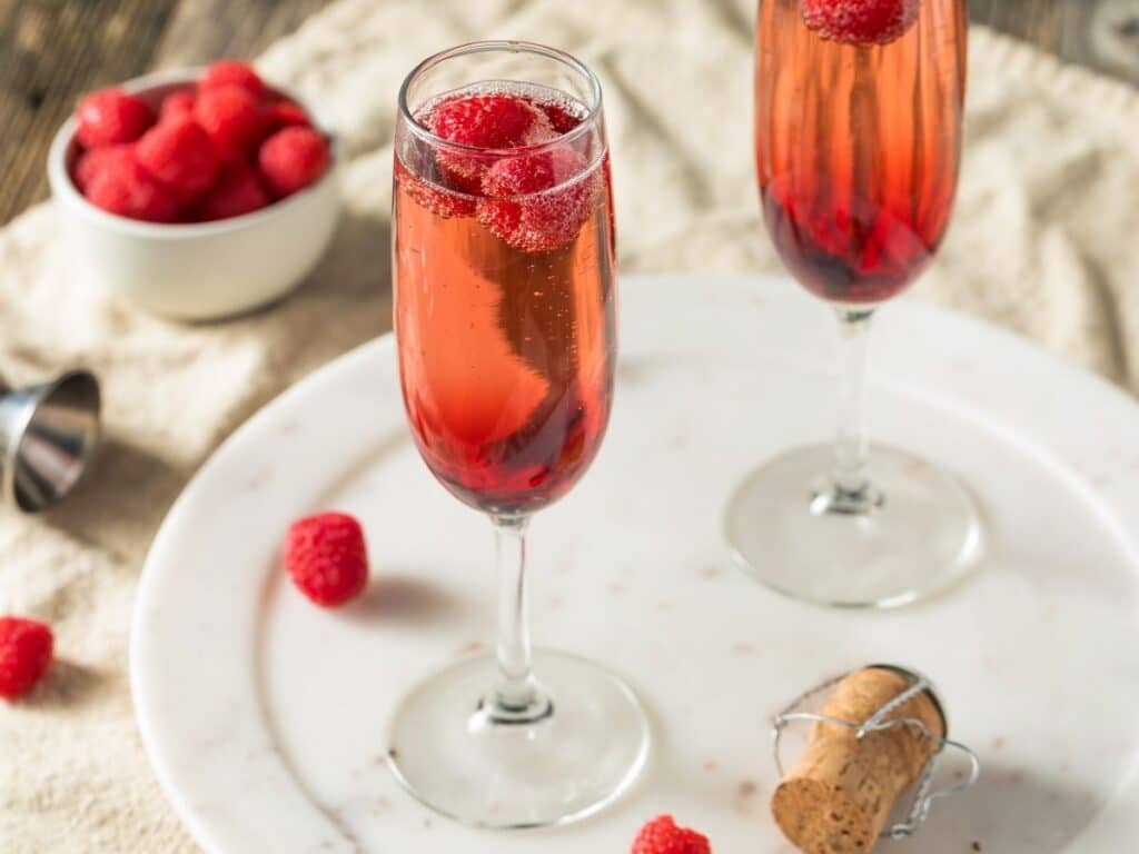 Two tall glasses of Kir Royale cocktail with floating raspberries, placed on a white marble tray with a bowl of fresh raspberries and a cork beside them.