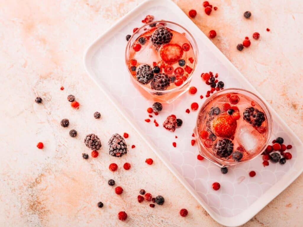 Two glasses of Lillet aperitif with ice and mixed berries, served on a white tray with scattered frozen berries around.