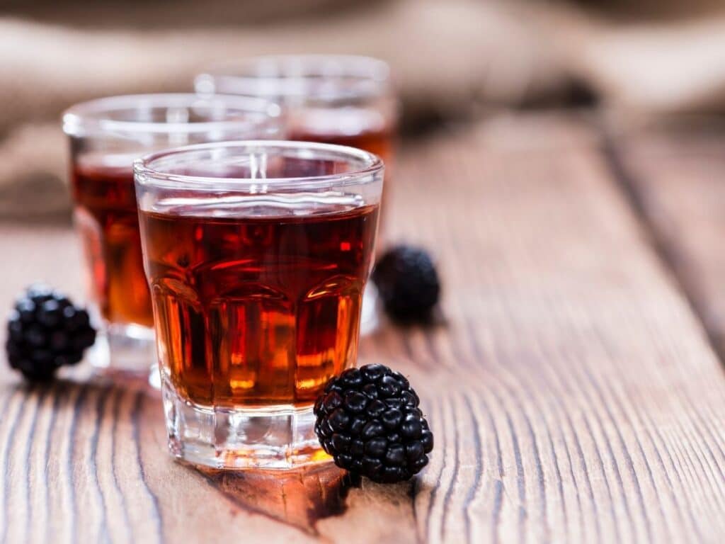 Three shot glasses of blackberry liqueur, garnished with fresh blackberries, set on a wooden surface.