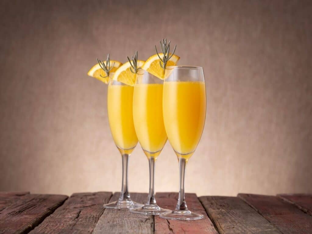 Three glasses of mimosa garnished with orange slices and rosemary sprigs, displayed on a rustic wooden table.