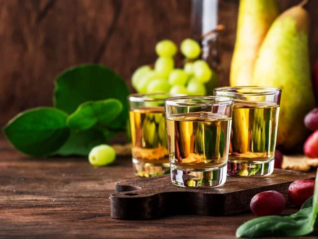 Three shot glasses of Ratafia de Champagne, surrounded by fresh grapes and pears, set on a wooden surface.