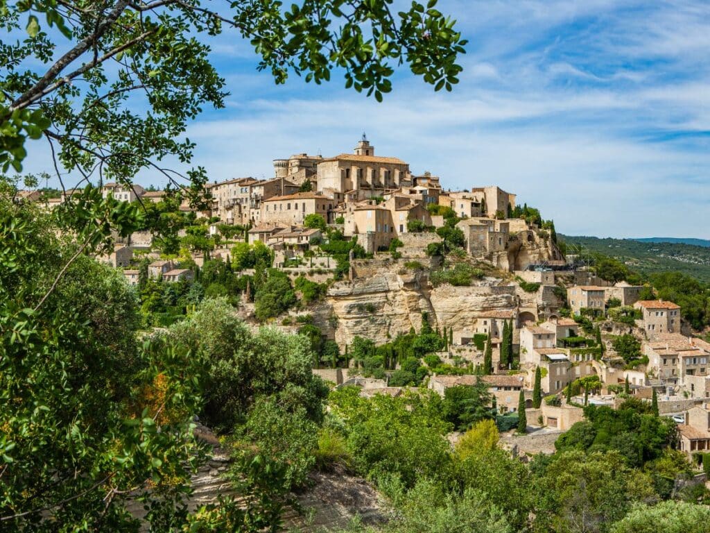 A historic hilltop village with stone buildings and greenery, seen through the framing branches of trees.