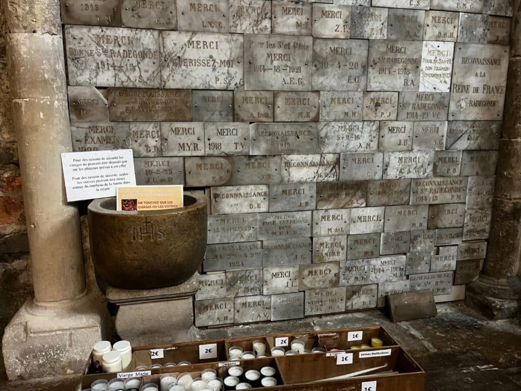 A wall covered with ex-voto plaques expressing gratitude to Sainte Radegonde, with a stone font below and labeled candles for sale.