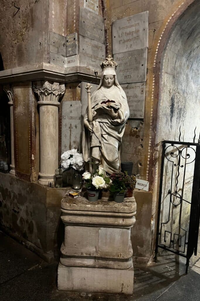A statue of Sainte Radegonde holding a book, surrounded by white and pink flowers in vases, set against a wall with numerous plaques in a stone crypt.