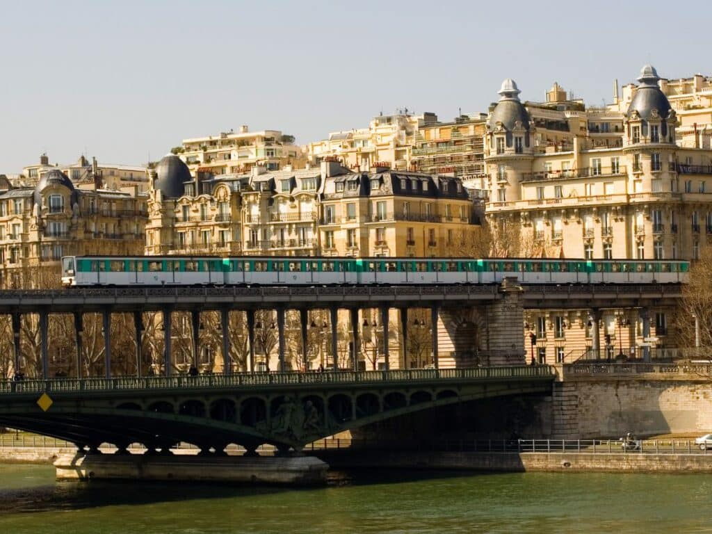 A train travels across a bridge in front of classic Parisian architecture with a green and white metro train passing over the Seine River.