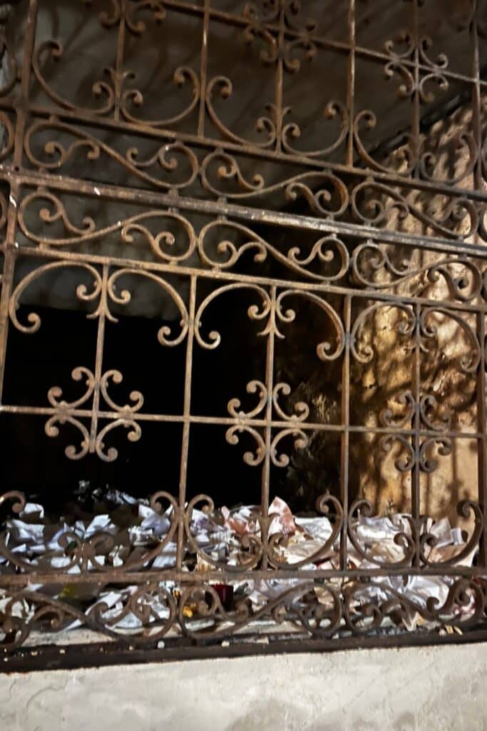 View through an ornate wrought iron gate into a crypt filled with folded papers and small offerings scattered on the floor.