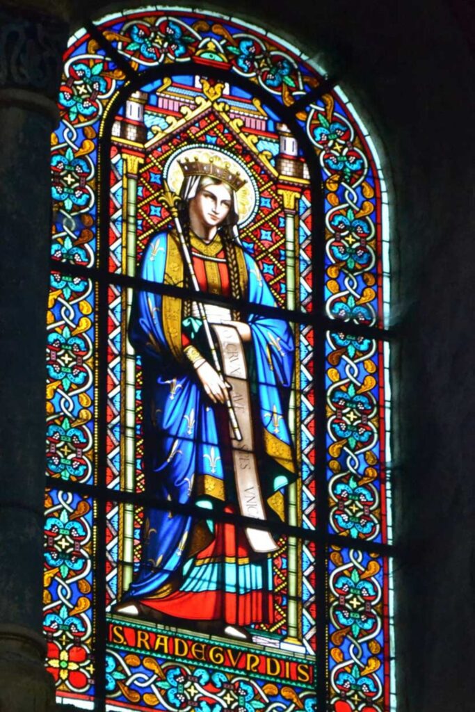 Stained glass window depicting Sainte Radegonde, dressed in royal blue and red garments, holding a scroll, with intricate floral and geometric patterns framing the image.