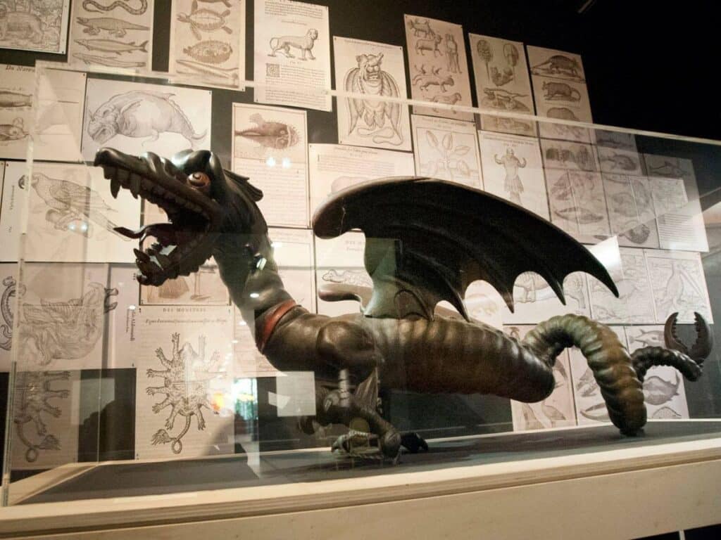 A model of La Grand'Goule, a dragon-like creature with wings and an open mouth, displayed in a museum exhibit with historical illustrations of mythical creatures on the wall behind it.