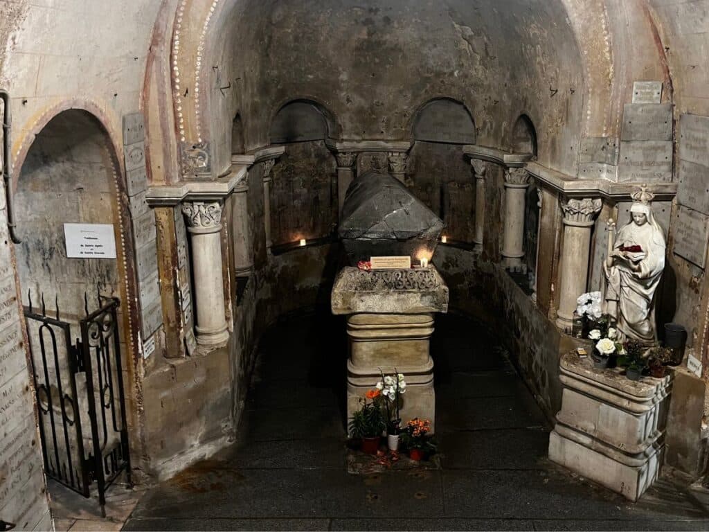 Interior of the tomb of Sainte Radegonde in Poitiers, France. The tomb is surrounded by ancient stone arches and columns, with a statue of Sainte Radegonde holding a book to the right, and various flowers and candles placed around the tombstone.
