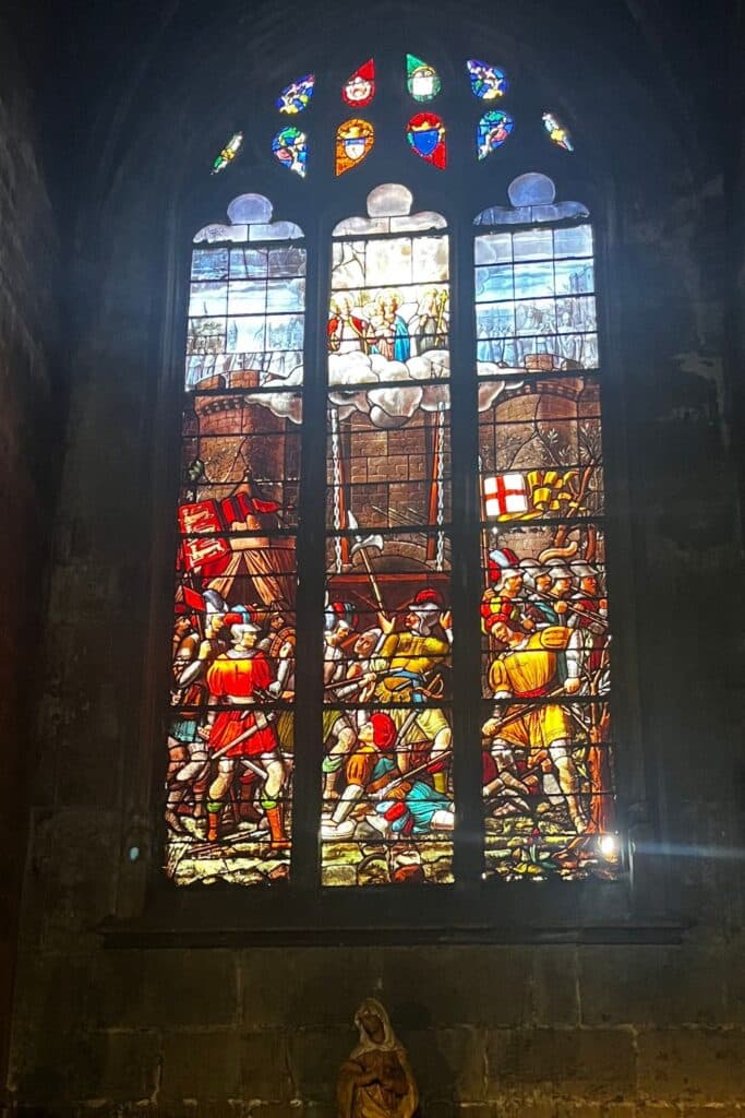 Stained glass window depicting 'The Miracle of the Keys,' showing a historical scene with soldiers in colorful armor at the bottom and a heavenly scene with angels at the top, framed by smaller shield emblems at the top of the window.