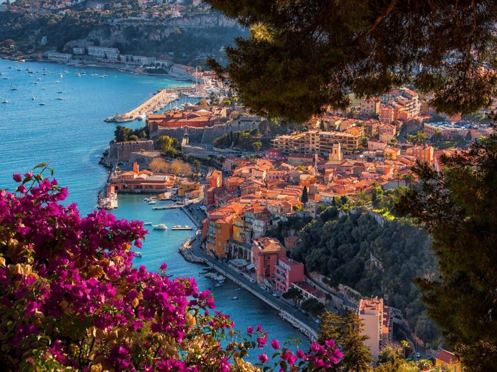 The coastal town of Villefranche-sur-Mer, overlooking the Mediterranean Sea with colorful houses cascading down the hillside and vibrant flowers in the foreground.