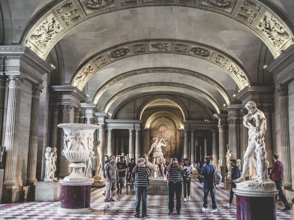 Visitors explore a grand, ornate hall in a museum in France, surrounded by classical sculptures and intricate architectural details.
