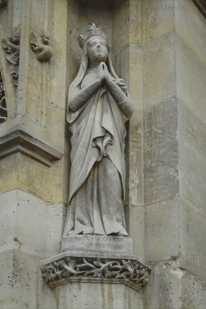A stone statue of Sainte Radegonde, depicted with a crown and clasped hands in a prayer position, set against the exterior wall of a historical building.