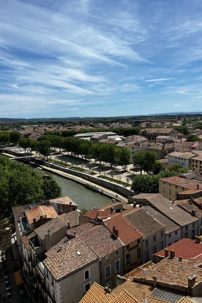 Aerial view from the top of the Donjon Gilles Aycelin, overlooking the city of Narbonne with its red-tiled roofs, a canal lined with trees, and a vast landscape under a bright blue sky.