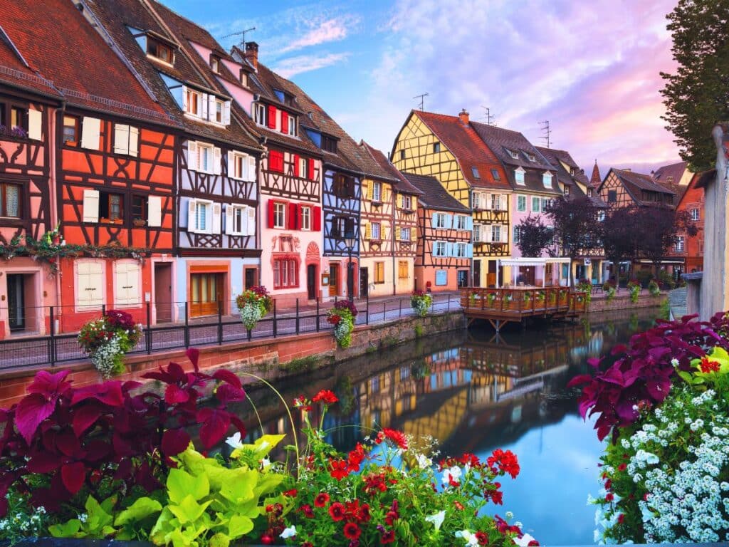 A vibrant canal-side street in an Alsatian village, with colorful half-timbered houses reflecting in the still water. Bright flowers in planters line the waterway, set against a backdrop of a pastel-colored sunset sky, evoking the region’s idyllic beauty.