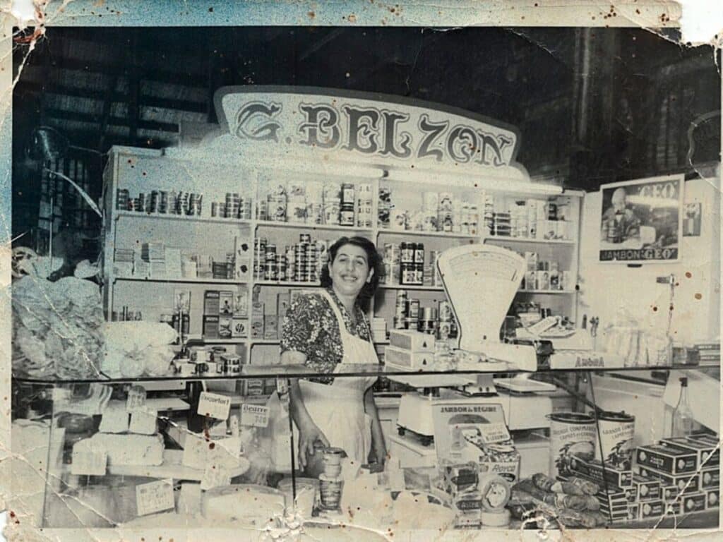 An old black-and-white photo of a woman standing behind the counter at a cheese and grocery shop named "Belzon." Shelves are lined with various canned goods and grocery items. The woman smiles as she prepares to serve customers.