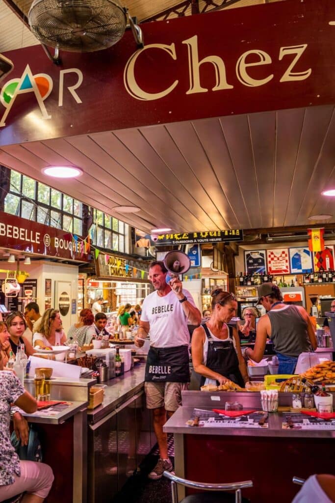 A bustling market scene at Chez Bebelle, where a man with a megaphone stands in front of a counter, interacting with customers. People are seated around the counter eating, while others work behind it, with colorful signage overhead.