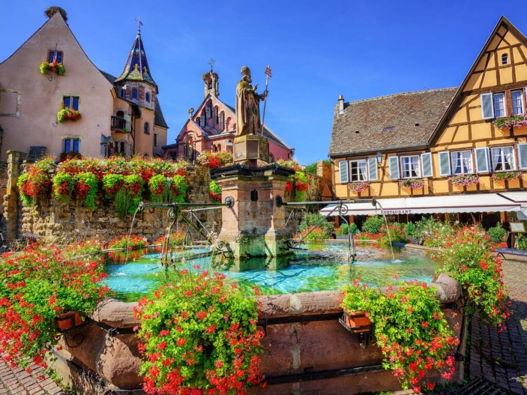 A charming square in Eguisheim, Alsace, featuring a stone fountain surrounded by beautifully preserved half-timbered houses. The houses are adorned with flower boxes overflowing with vibrant blooms, creating a fairytale atmosphere beneath a bright blue sky.