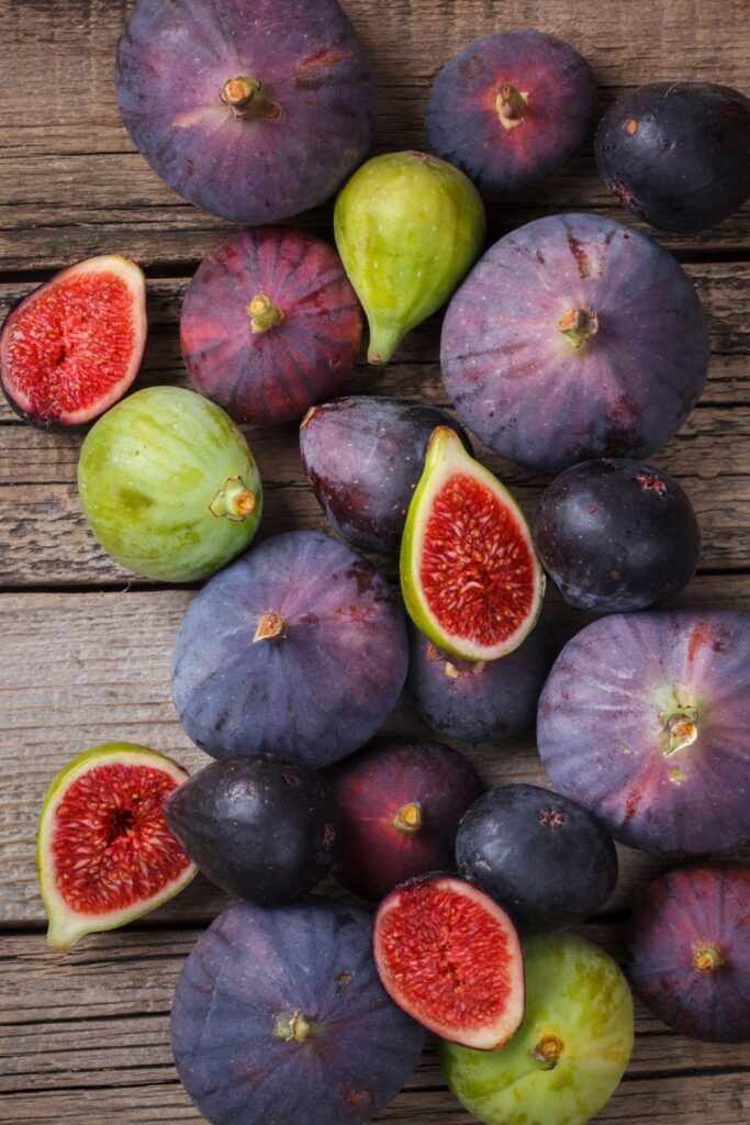 A variety of fresh figs spread across a wooden surface, showcasing different colors and sizes, including deep purple, green, and sliced red figs.