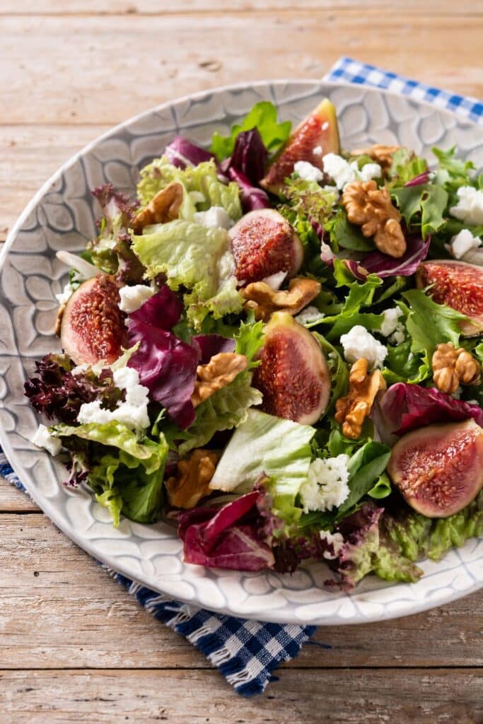 A vibrant salad of fresh figs, mixed greens, crumbled feta, and walnuts, arranged in a decorative bowl on a rustic wooden table.