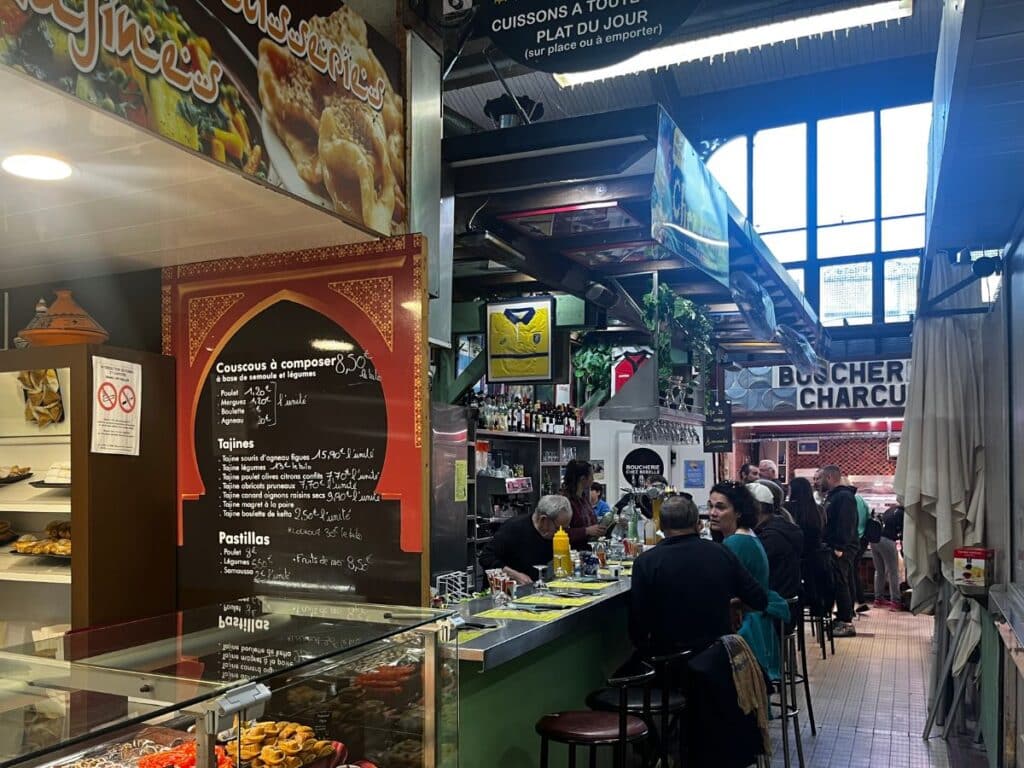 A lively market stall featuring Moroccan-style cuisine, with a sign listing menu items like couscous and tajines. Customers sit at a bar counter, engaging in conversation, with a butcher shop visible further inside the market.