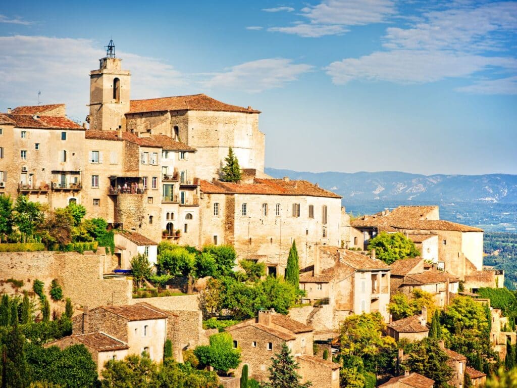 The hilltop village of Gordes, featuring stone buildings with terracotta roofs, surrounded by lush greenery and overlooking a valley.