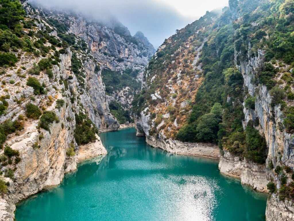 The turquoise waters of the Gorges du Verdon canyon, flanked by steep rocky cliffs and dense green foliage.