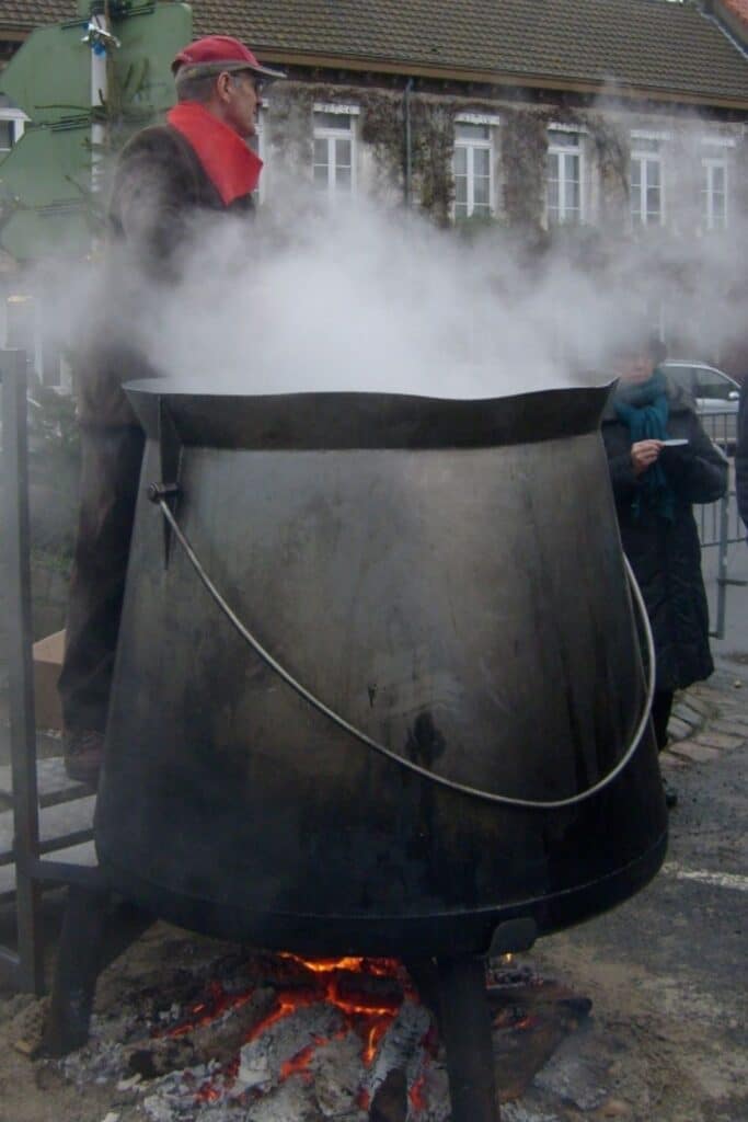 An elderly man wearing a red scarf and hat stands beside a large, steaming cauldron suspended over a wood fire, with a few people in the background on a village street.