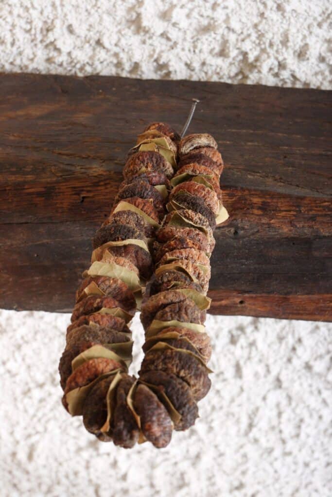 A strand of dried figs threaded together with bay leaves, hanging from a wooden beam against a textured white wall.