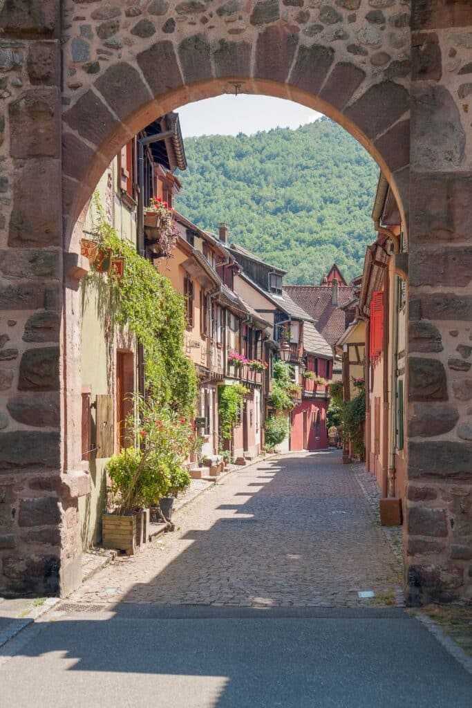 A cobblestone street in Kaysersberg, framed by a stone archway, leading to charming half-timbered houses covered in greenery. The village is nestled against a lush green hill, evoking a serene, historic ambiance.