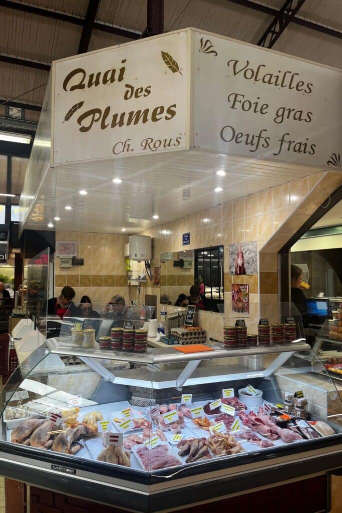 A display case filled with fresh cuts of poultry, foie gras, and other meats at a butcher stall named "Quai des Plumes." The sign above highlights the specialty products, and several people are seen working behind the counter.