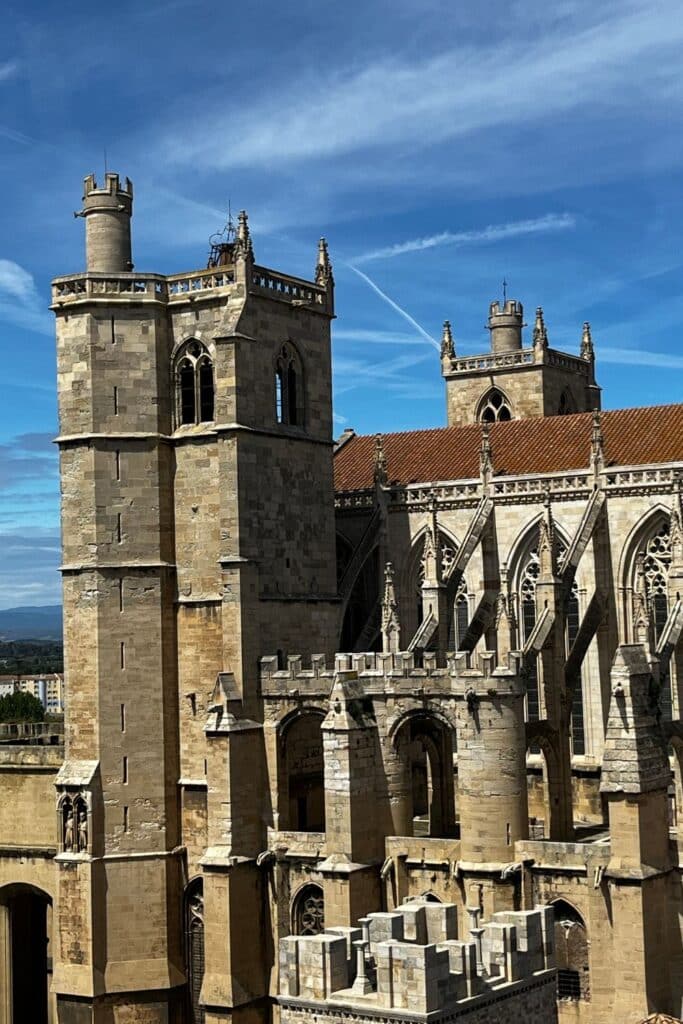 Close-up view of Narbonne Cathedral with its towering spires, detailed stone arches, and medieval architecture set against a backdrop of blue sky and wispy clouds.