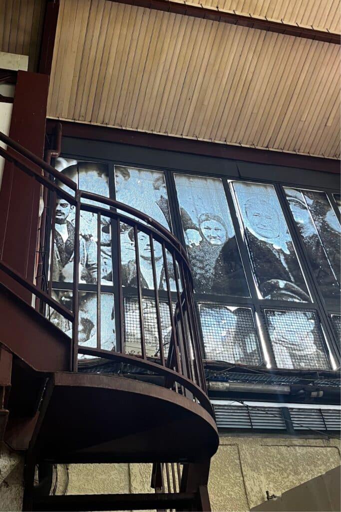 A spiral staircase in an old market building, leading up to large windows covered in faded, historical photos of people. The vintage Belle Époque design of the market is highlighted by its iron structure and weathered aesthetic.