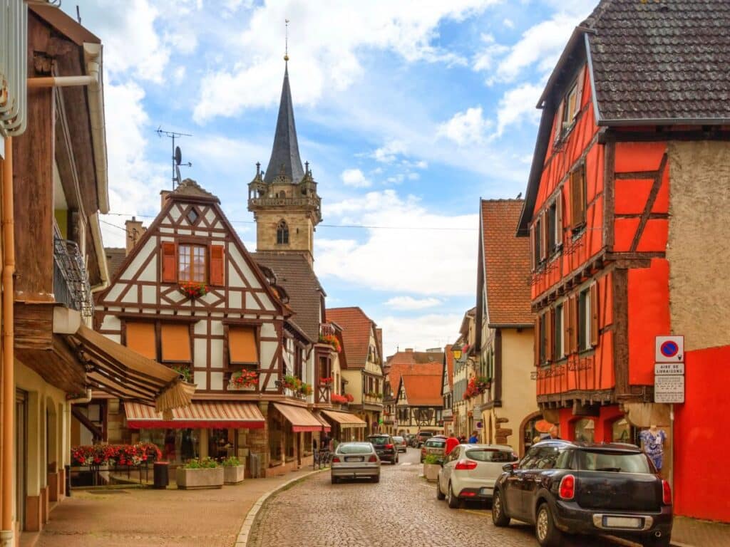 A bustling street in Obernai, lined with colorful half-timbered houses and featuring a church steeple in the background. Flower boxes and outdoor seating add to the lively yet quaint atmosphere of this Alsatian village.