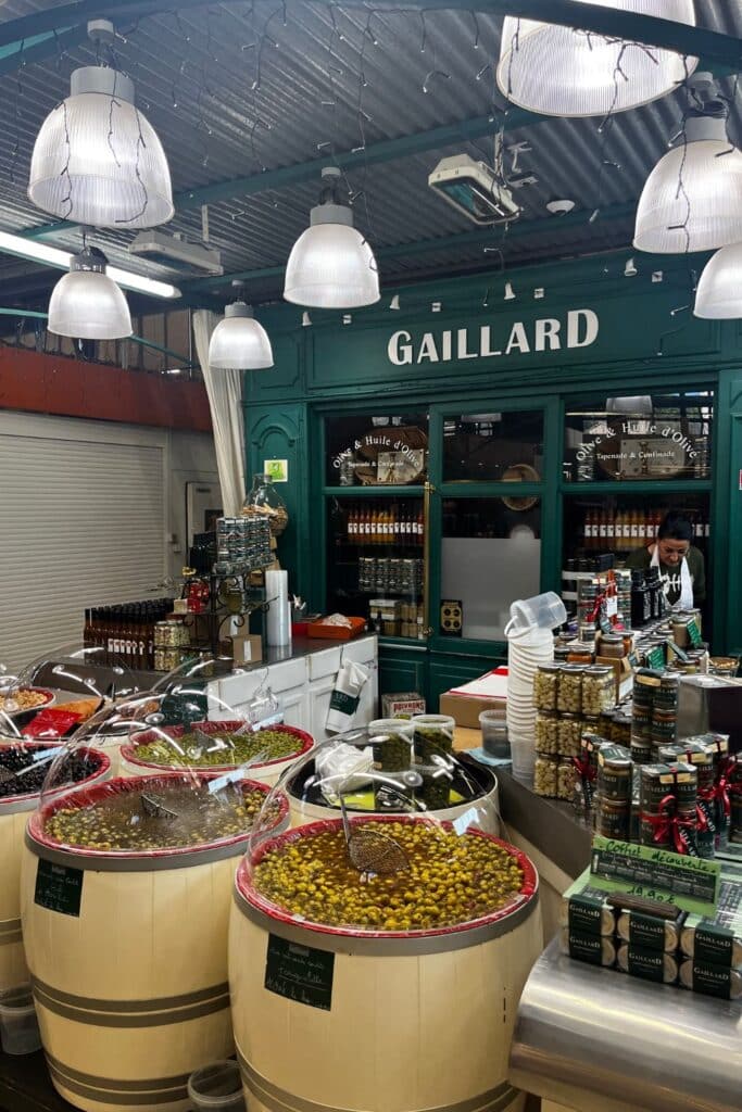 A market stall named "Gaillard" featuring large barrels filled with different varieties of olives and pickled vegetables. The stall is decorated with hanging lights and jars of olive oil and tapenade are displayed on the counter.