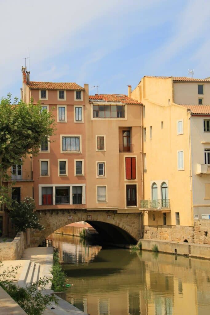 A row of pastel-colored buildings along a canal in Narbonne, France, with reflections in the water and a stone bridge, highlighting the city’s quaint and historic atmosphere.