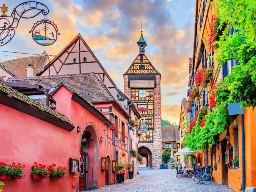 A picturesque view of Riquewihr’s narrow street lined with brightly painted half-timbered houses adorned with flower boxes. A tall clock tower stands at the end of the street, adding to the village’s medieval charm under a vibrant sunset sky.