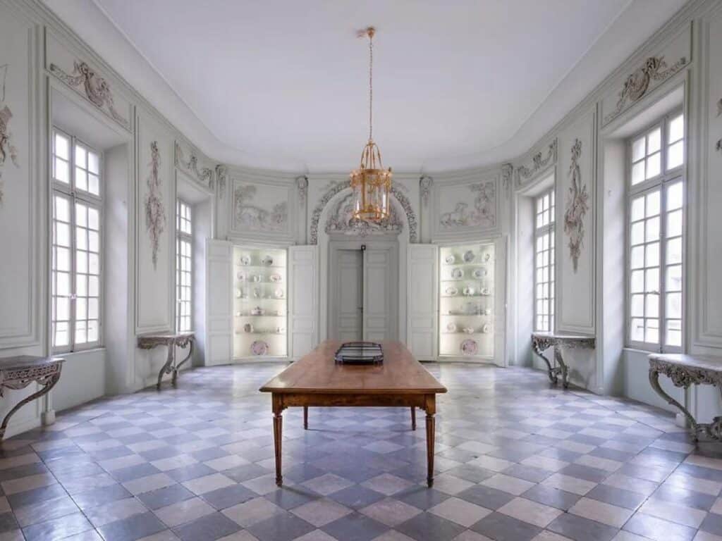Elegant dining room with a long wooden table, ornate white walls with intricate moldings, large windows, and a chandelier hanging from the ceiling.