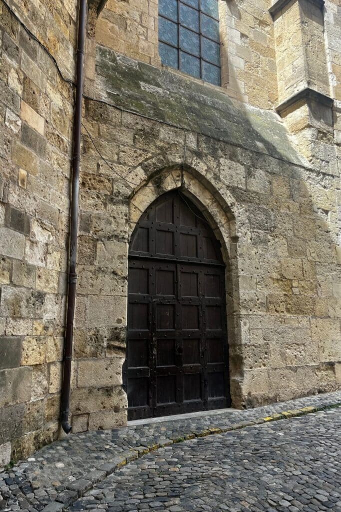 A dark wooden door set into the stone wall of a medieval church in Narbonne, France, with a cobblestone pathway in front, emphasizing the city’s historical architecture.