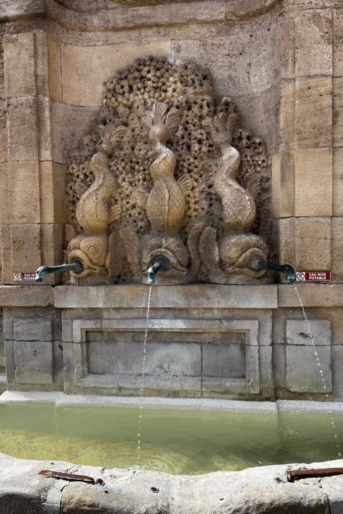 A historical stone fountain in Narbonne, France, featuring intricately carved fish sculptures with water spouts flowing into a basin. The detailed artistry reflects Narbonne’s rich cultural heritage.