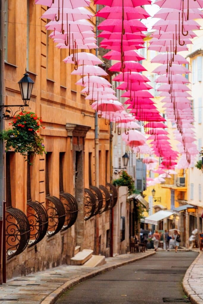 A charming narrow street in Grasse, lined with pastel-colored buildings, featuring a whimsical overhead display of pink umbrellas hanging above, creating a vibrant and playful atmosphere.