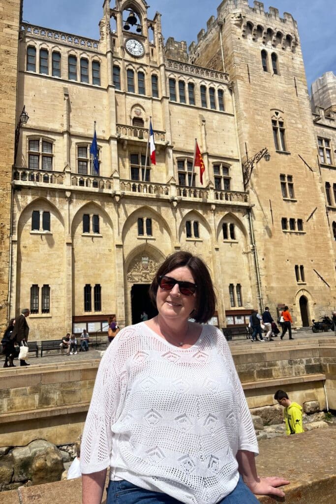 A woman posing in front of the historic Donjon Gilles Aycelin in Narbonne, with its ornate stone facade, clock tower, and flags.